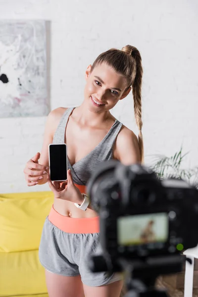 Selektiver Fokus der fröhlichen Sportlerin, die ihr Smartphone mit leerem Bildschirm in der Nähe der Digitalkamera im Wohnzimmer hält — Stockfoto