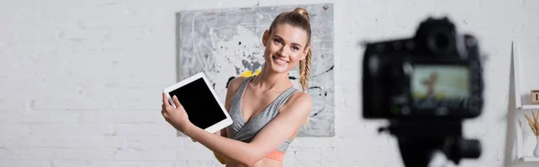 Panoramaaufnahme einer fröhlichen Sportlerin, die ein digitales Tablet in der Nähe der Digitalkamera im Wohnzimmer hält — Stockfoto