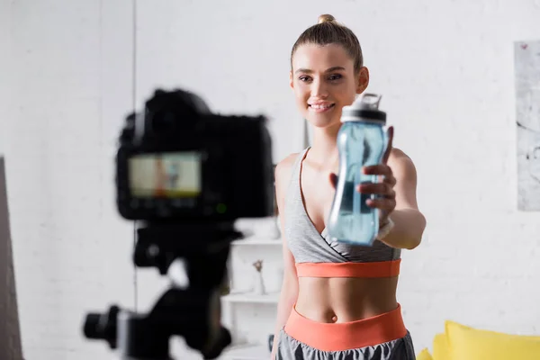 Selective focus of smiling sportswoman showing sports bottle at digital camera in living room — Stock Photo