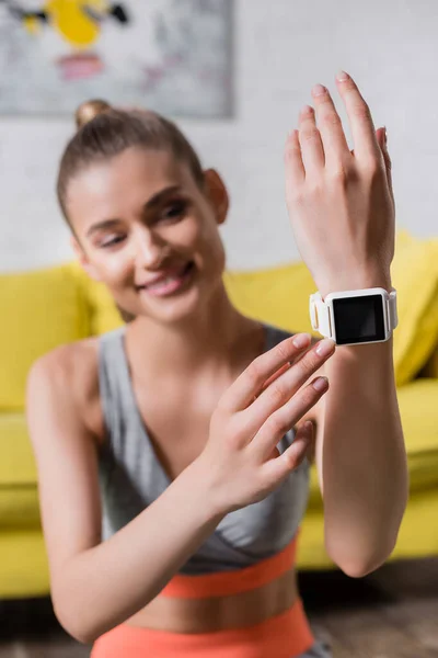 Selective focus of smiling sportswoman touching smartwatch at home — Stock Photo