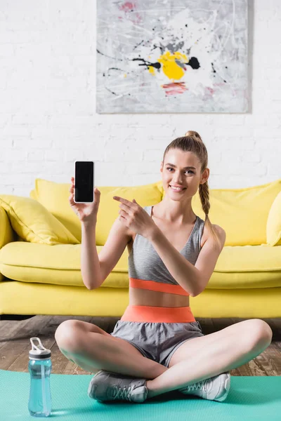 Desportista sorridente apontando para o smartphone com tela em branco enquanto sentado no tapete de fitness em casa — Fotografia de Stock