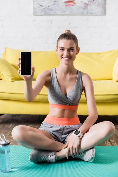 Позитивна спортсменка, що показує смартфон на камеру на фітнес-маті вдома — стокове фото