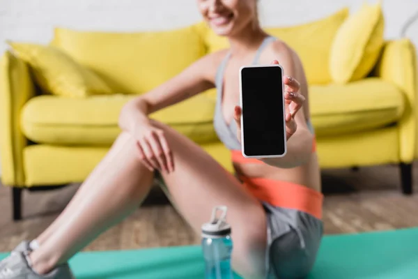 Обрізаний вид усміхненої спортсменки, що показує смартфон з порожнім екраном біля спортивної пляшки на фітнес-маті — стокове фото