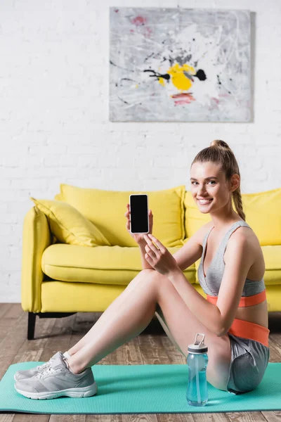 Sportive positive tenant smartphone avec écran blanc près de la bouteille de sport sur tapis de fitness — Photo de stock