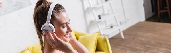 Foto panoramica di una giovane donna sorridente mentre ascolta musica in cuffia a casa — Foto stock