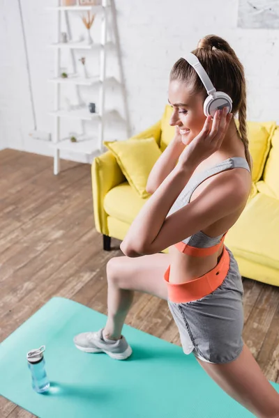 Foco seletivo de esportista sorridente em fones de ouvido trabalhando no tapete de fitness na sala de estar — Fotografia de Stock