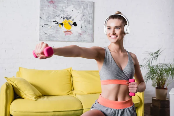 Focus selettivo della sportiva sorridente in allenamento cuffie con manubri in soggiorno — Foto stock