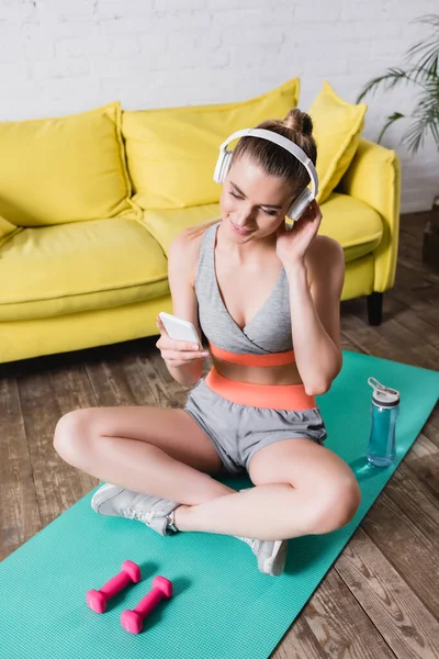 Sportswoman sorrindo em fones de ouvido usando smartphone enquanto sentado no tapete de fitness em casa — Fotografia de Stock