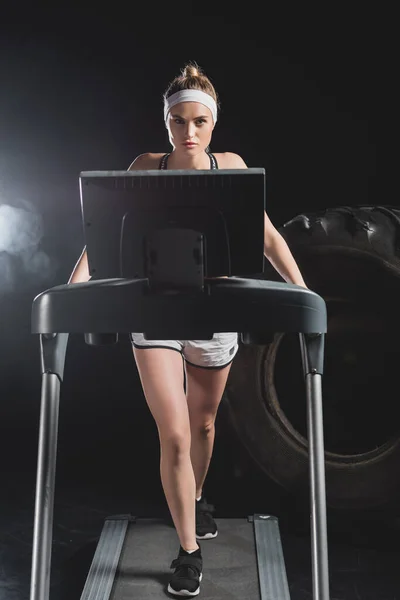 Selective focus of young sportswoman training on treadmill near tire in gym with smoke — Stock Photo