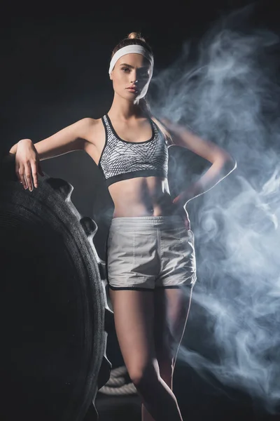 Sportswoman with hand on hip looking at camera near tire in gym with smoke — Stock Photo