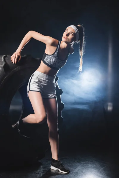 Sportswoman en vêtements de sport et bandeau exercice avec pneu pendant l'entraînement dans la salle de gym avec de la fumée — Photo de stock