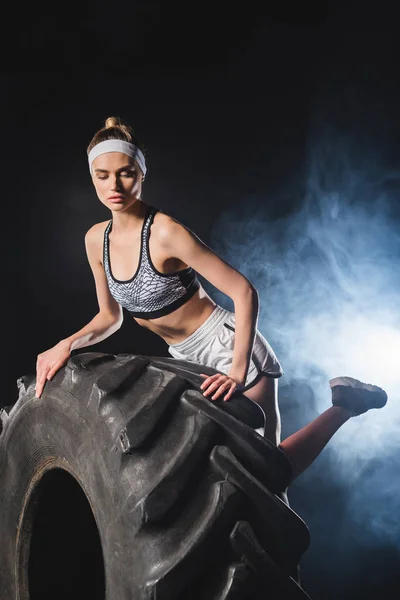 Selective focus of young sportswoman touching tire in gym with smoke — Stock Photo