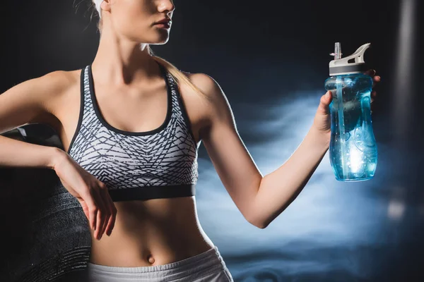 Cropped view of sportswoman holding sports bottle near tire in gym with smoke — Stock Photo