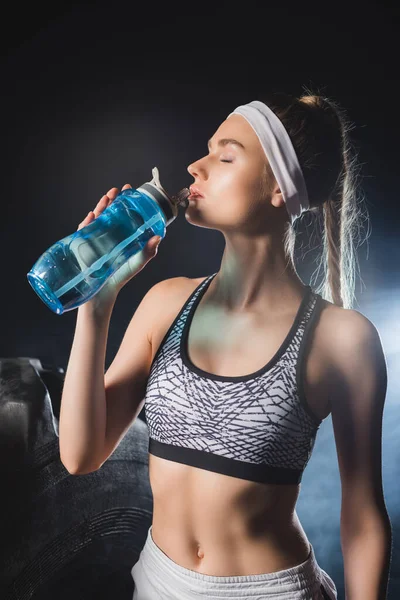 Sportswoman drinking water near tire in gym with smoke — Stock Photo