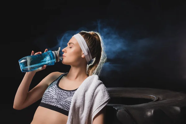 Joven deportista con toalla bebiendo agua cerca de neumático en gimnasio con humo - foto de stock