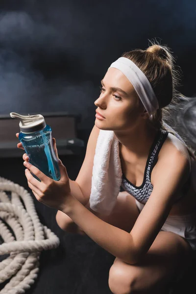 Selektiver Fokus der Sportlerin mit Handtuch, die Sportflasche in der Nähe von Kampfseil in der Sporthalle mit Rauch hält — Stockfoto