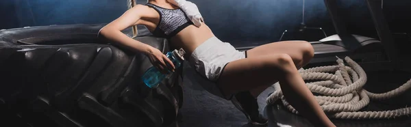 Récolte panoramique de sportive avec bouteille de sport et serviette près des pneus et corde de combat dans la salle de gym avec de la fumée — Photo de stock