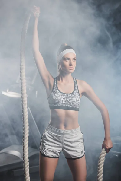 Selective focus of young sportswoman holding battle rope in gym with smoke — Stock Photo