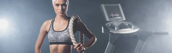 Panoramic shot of sportswoman looking at camera while holding battle rope near treadmill in gym with smoke — Stock Photo