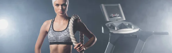 Prise de vue panoramique d'une sportive regardant une caméra tout en tenant une corde de combat près d'un tapis roulant dans un gymnase avec de la fumée — Photo de stock