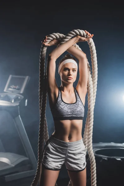 Young sportswoman looking at camera while holding battle rope in sports center with smoke — Stock Photo