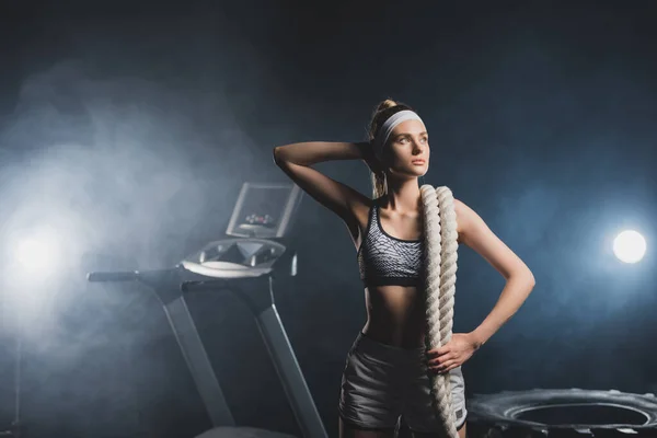Young sportswoman with battle rope looking away near tire and treadmill in gym with smoke — Stock Photo