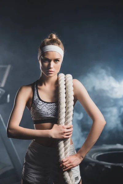 Young sportswoman with battle rope looking at camera in sports center with smoke — Stock Photo