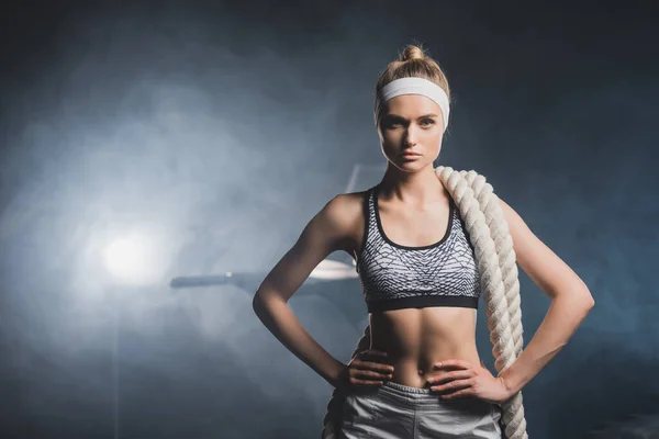 Sportswoman in headband with battle rope looking at camera in gym with smoke — Stock Photo