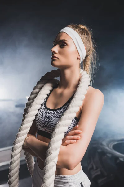 Joven deportista con brazos cruzados y cuerda de batalla de pie en el gimnasio con humo - foto de stock