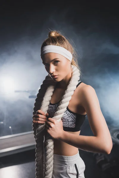 Mujer joven en ropa deportiva y diadema sosteniendo la cuerda de batalla mientras mira la cámara en el gimnasio con humo - foto de stock