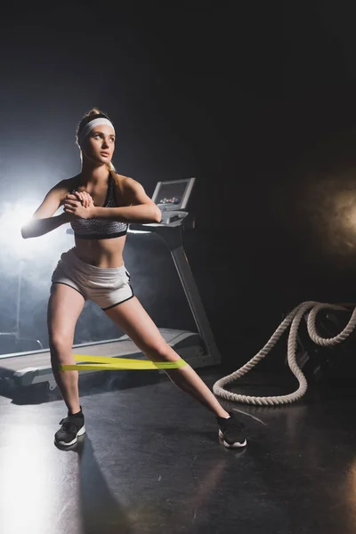 Young sportswoman exercising with resistance band near battle rope and treadmill in gym with smoke — Stock Photo