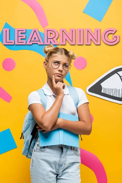 Nachdenkliches Schulmädchen mit Brille schaut weg und hält Buch neben Bleistift, bunten Elementen und gelbem Schriftzug — Stockfoto
