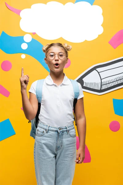 Colegiala sorprendida en gafas que muestran idea signo cerca de burbuja pensamiento, lápiz de papel y elementos abstractos en amarillo - foto de stock