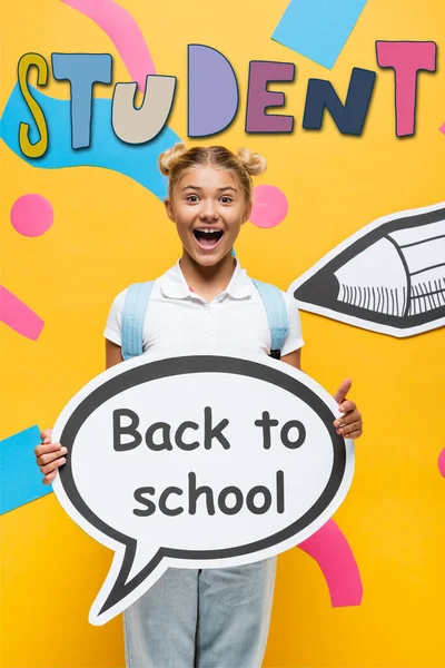 Excited schoolchild holding speech bubble with back to school lettering near paper elements and pencil on yellow — Stock Photo