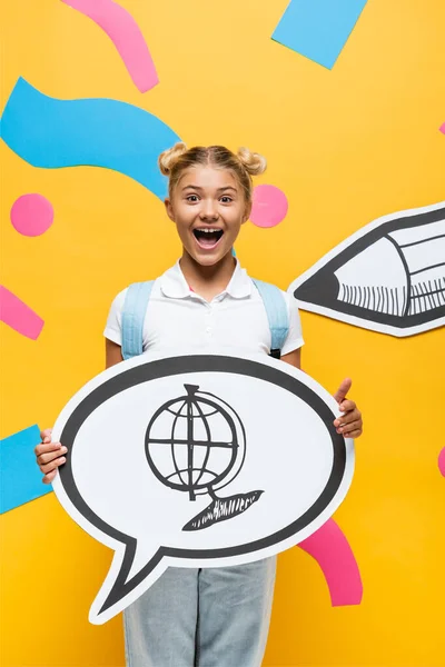 Écolier excité tenant bulle de parole avec illustration globe près des éléments en papier et crayon sur jaune — Photo de stock