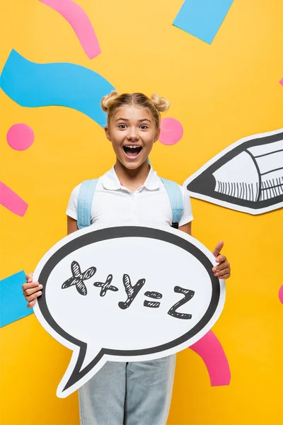 Aufgeregtes Schulkind hält Sprechblase mit mathematischer Formel-Illustration in der Nähe von Papierelementen und Bleistift auf gelb — Stockfoto