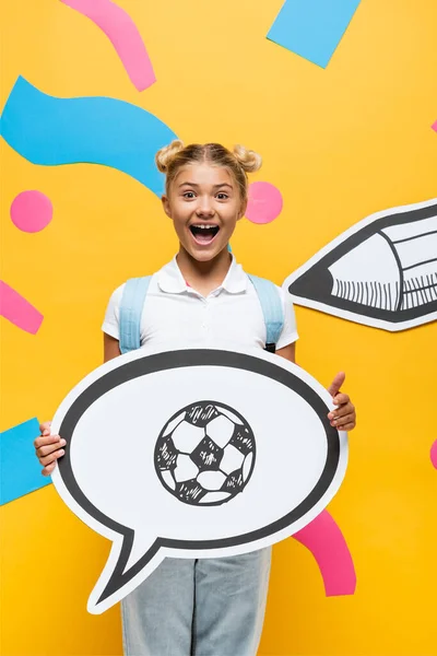 Écolier excité tenant bulle de parole avec illustration de football près des éléments de papier et crayon sur jaune — Photo de stock