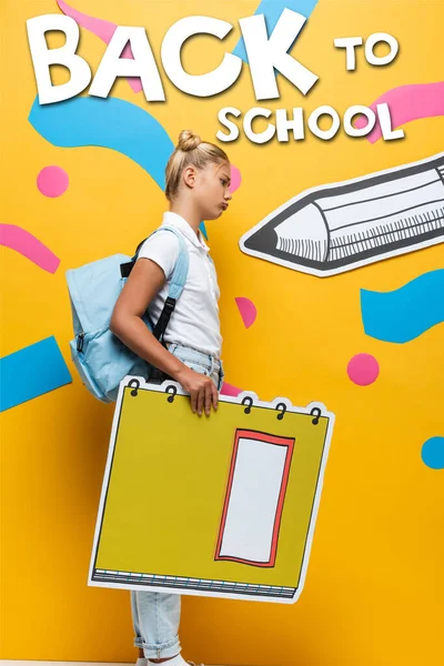 Seitenansicht eines aufgebrachten Schulmädchens mit Notizbuch-Maquette in der Nähe von Schulbuchstaben, Bleistift und dekorativen Elementen auf gelb — Stockfoto