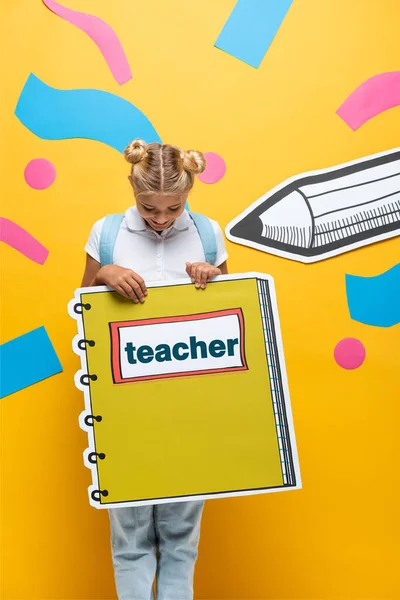 Joyful schoolkid holding copy book maquette with teacher lettering near paper cut pencil and decorative elements on yellow — Stock Photo