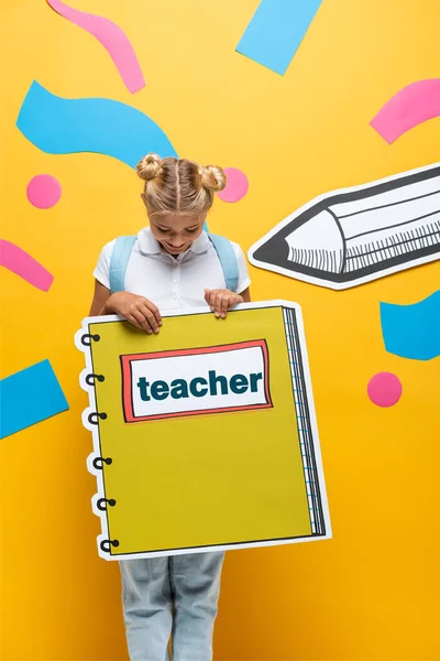 Alegre colegial sosteniendo maqueta libro de copia con letras de profesor cerca de papel lápiz cortado y elementos decorativos en amarillo - foto de stock