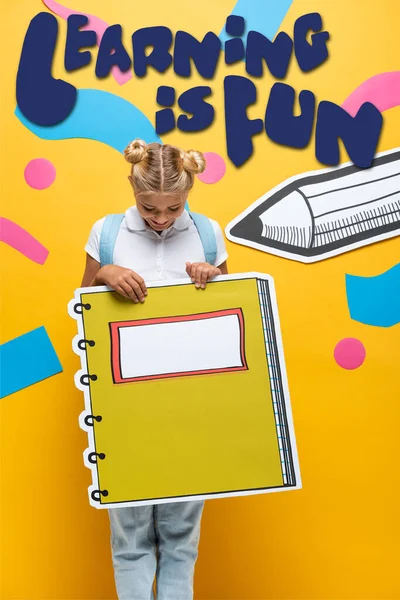 Aluna alegre segurando maquete livro de cópia perto de aprendizagem é divertido lettering, lápis de corte de papel e elementos decorativos em amarelo — Fotografia de Stock