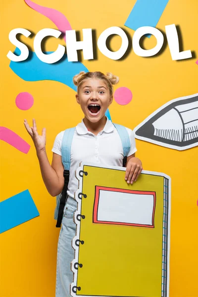 Écolière excitée avec maquette cahier geste près des éléments décoratifs, lettrage de l'école et crayon de coupe de papier sur jaune — Photo de stock