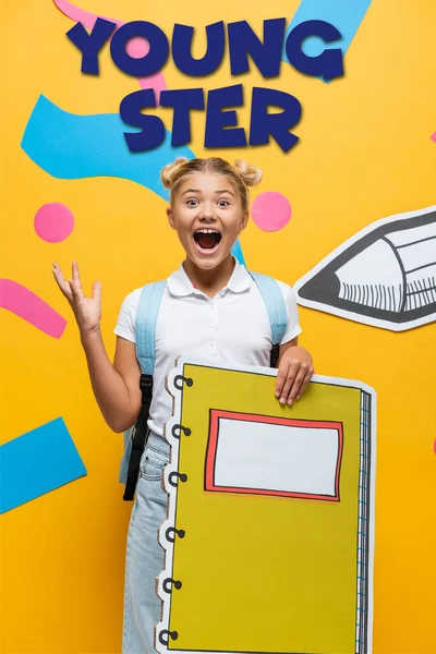 Écolière excitée avec maquette cahier geste près des éléments décoratifs, lettrage des jeunes et crayon de papier découpé sur jaune — Photo de stock