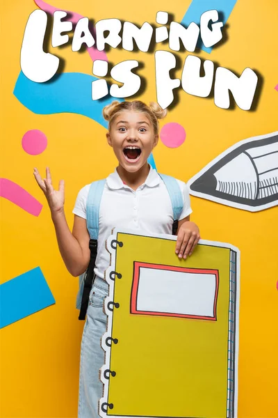 Excited schoolgirl with notebook maquette gesturing near decorative elements, learning is fun lettering and paper cut pencil on yellow — Stock Photo