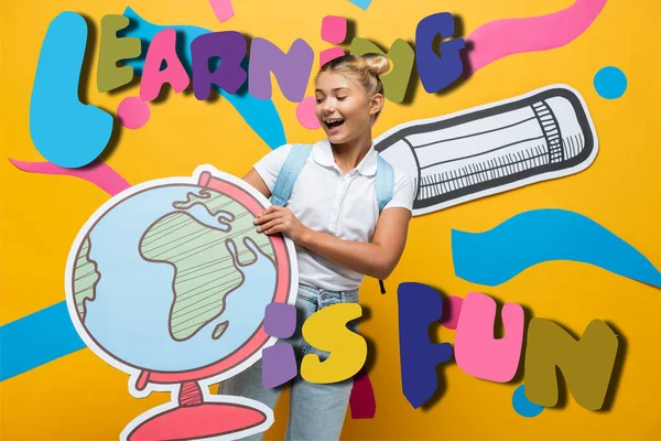 Alegre colegiala celebración globo maqueta cerca de aprender es divertido letras, lápiz de papel y elementos abstractos en amarillo - foto de stock