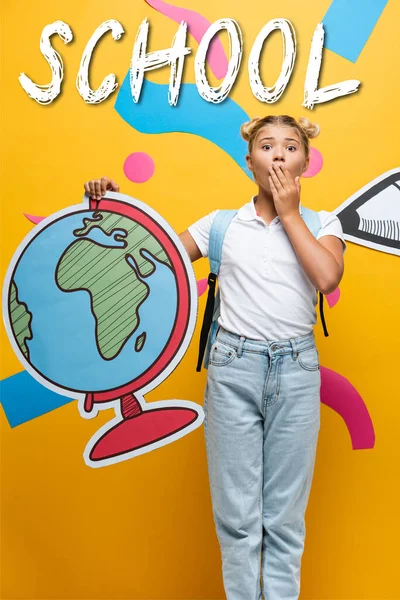 Shocked schoolgirl covering mouth while holding globe maquette near school lettering, paper pencil and decorative elements on yellow — Stock Photo