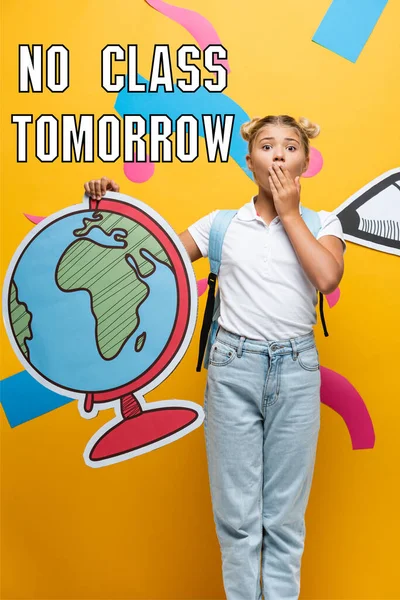 Shocked schoolgirl covering mouth while holding globe maquette near no class tomorrow lettering, paper pencil and decorative elements on yellow — Stock Photo