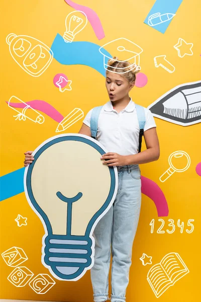 Amazed pupil holding light bubble maquette near multicolored elements, paper pencil and illustration on yellow — Stock Photo