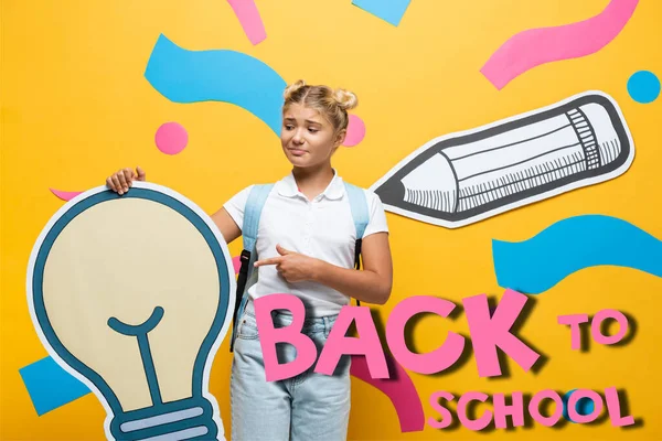 Confused schoolgirl pointing with finger at paper light bulb near decorative elements and back to school lettering on yellow — Stock Photo