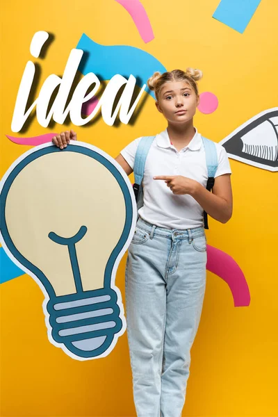 Schoolgirl with backpack pointing with finger at decorative light bulb near paper art and idea lettering on yellow background — Stock Photo